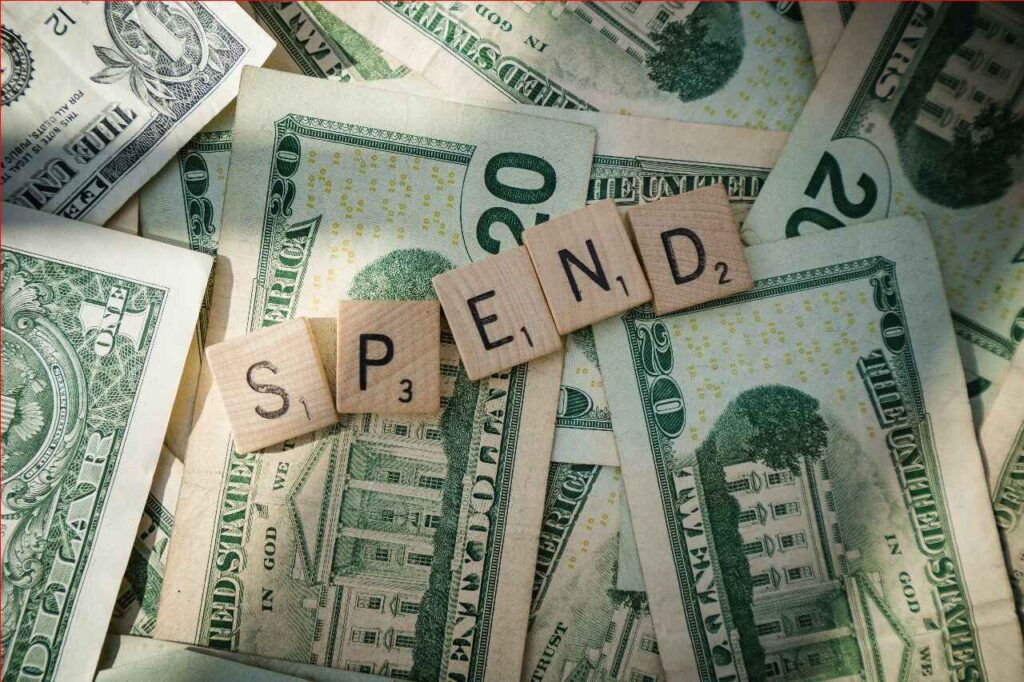Wooden Scrabble tiles spelling “SPEND” placed over U.S. dollar bills, representing everyday spending habits and how mindful choices can help you save money fast.