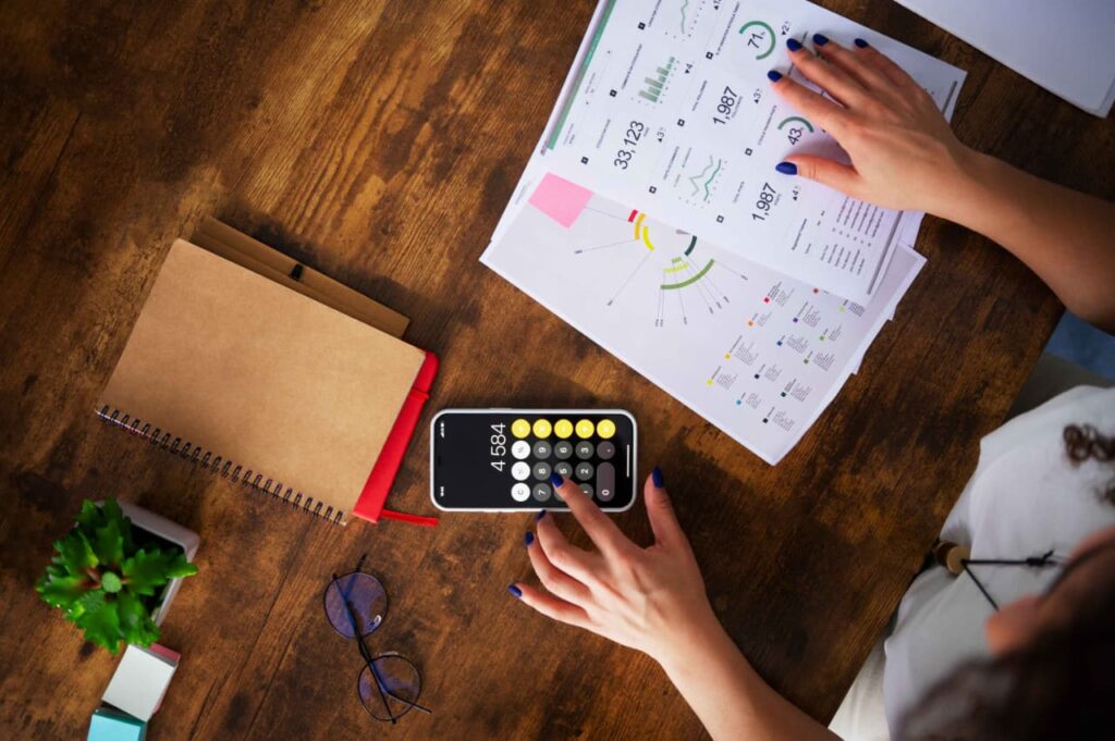 A person is working at a wooden desk with a smartphone displaying the number 4584 on a calculator app. The desk also holds a notebook, eyeglasses, a small potted plant, and several sheets of paper featuring graphs and charts. One hand is interacting with the phone while the other holds the papers, suggesting active engagement in data analysis or financial planning.